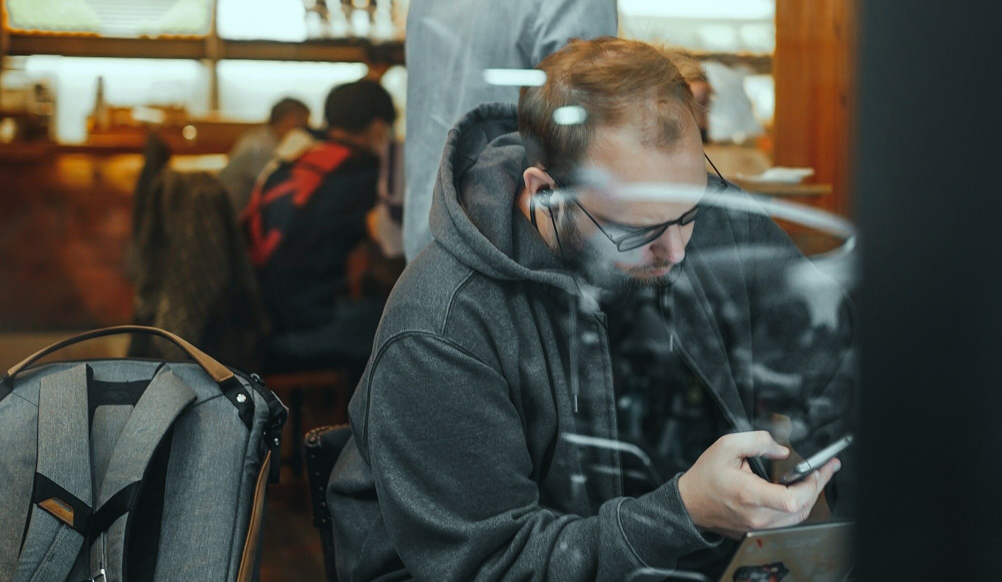 A man in a grey sweatshirt, wearing black ear buds, scrolls through his phone with his laptop open in front of him. On the chair to his left sits a grey and brown backpack. Behind him, the movement of what looks to be a busy cafe