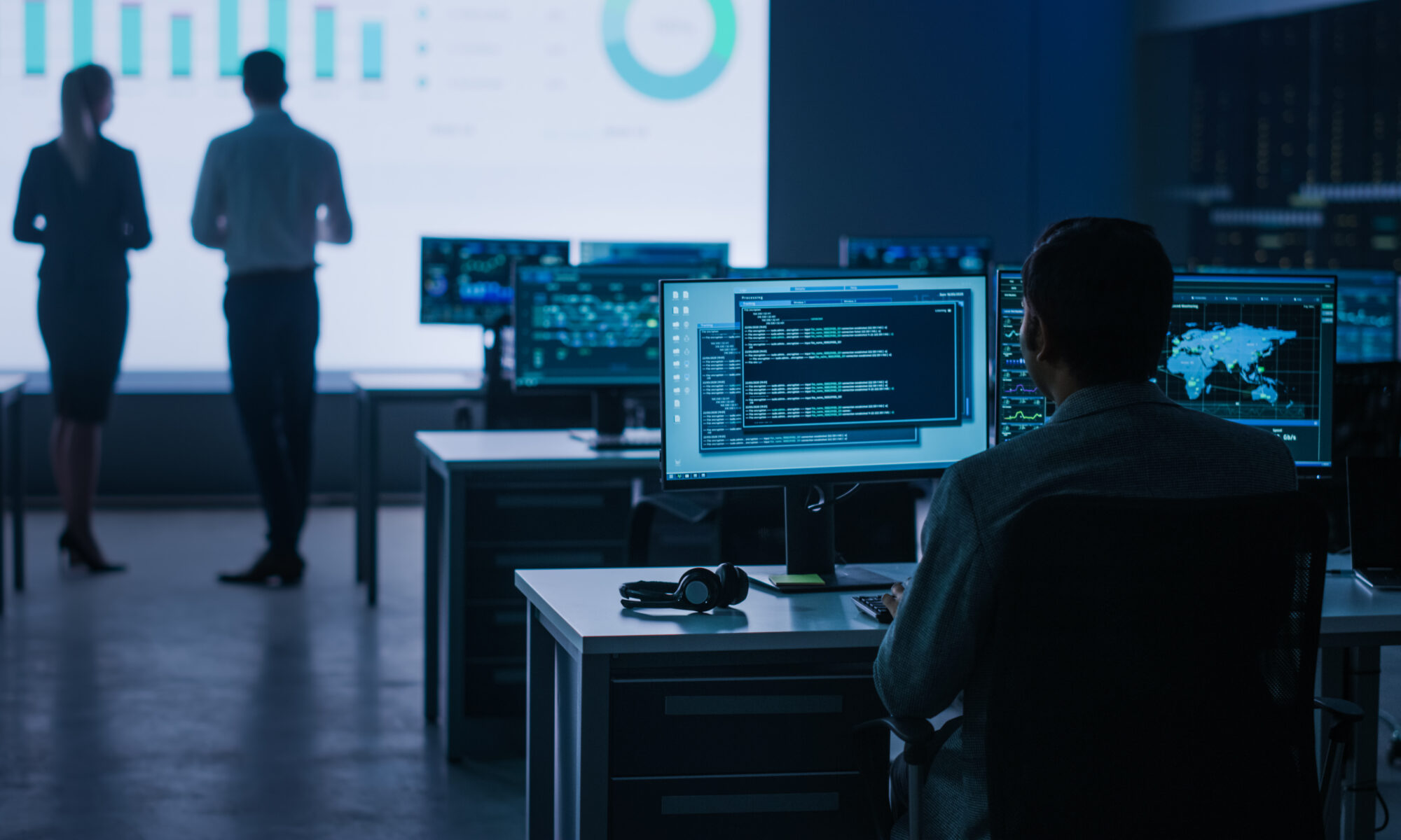 Back View Shot of a Professional Coder is Working on Personal Computer with Computer Codes, Infrastructure Infographics, Data and World Map. Telecommunications Control Room with Working Professionals.